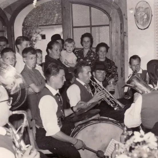 Ein Schwarz-Weiß-Foto zeigt eine Gruppe von Menschen, die drinnen versammelt sind und Musikinstrumente spielen. Die zentrale Figur hält eine Trompete, während andere Trommeln und Posaunen spielen. Eine Frau hält ein Kind und ein Mann trinkt aus einem Glas.