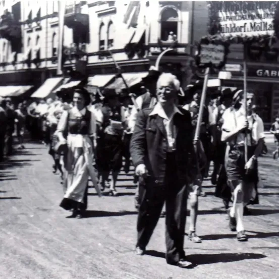 Ein historisches Foto zeigt einen älteren Mann, der in einer Parade auf einer Straße geht. Er trägt einen Mantel und hält einen Stock. Hinter ihm marschieren Menschen in einer Reihe.