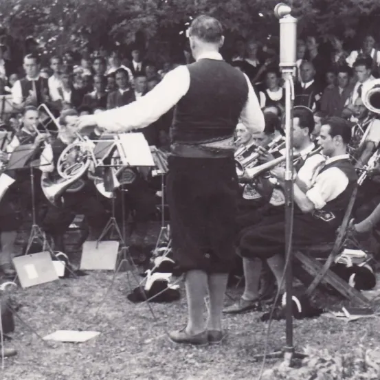 Ein Schwarz-Weiß-Foto einer Band, die vor einem Publikum mit Blechblasinstrumenten spielt. Ein Dirigent leitet die Band.