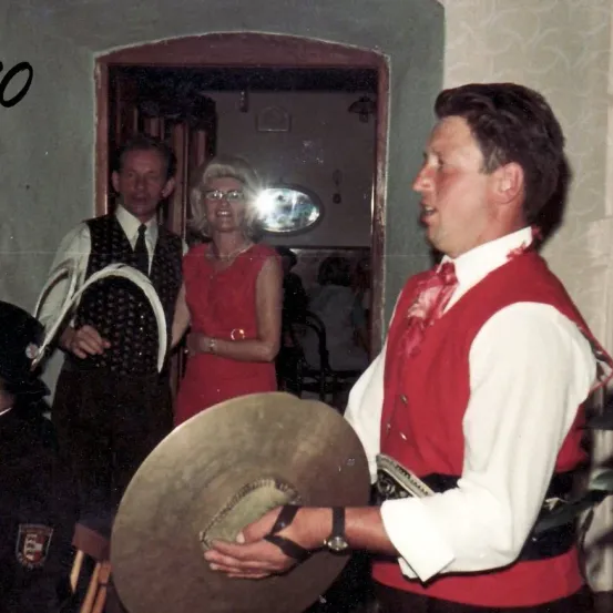 Ein Mann in einer roten Weste hält eine Zymbal, während ein Mann und eine Frau im Hintergrund zu sprechen scheinen.