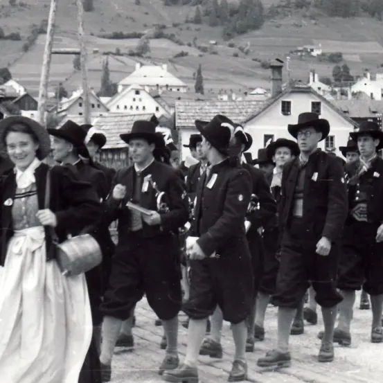 Eine Gruppe von Menschen in traditioneller Kleidung marschiert auf einer Straße. Sie tragen Hüte, Schuhe und einige tragen Gegenstände. Hinter ihnen befinden sich Gebäude und Hügel.