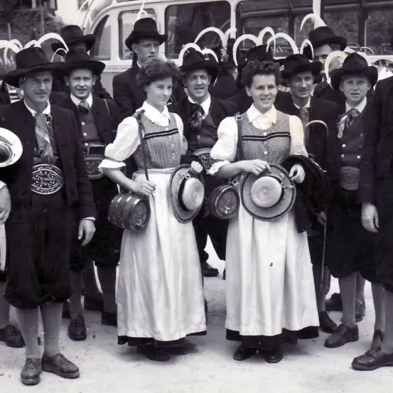 Ein Schwarz-Weiß-Foto zeigt eine Gruppe von Menschen in traditioneller deutscher Kleidung vor einem Bus. Einige tragen Hüte und Taschen. Eine Frau in der Mitte hält einen Hut.