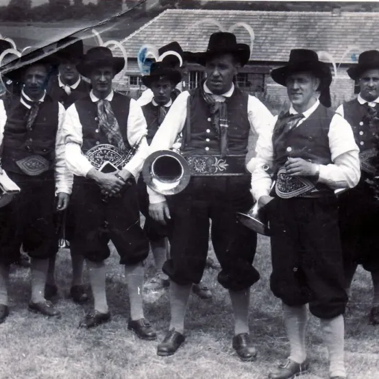 Eine Gruppe von Männern in traditioneller Kleidung mit Hüten und Schuhen steht auf Gras und hält Instrumente. Sie scheinen Mitglieder einer Blaskapelle zu sein, einer hält eine Trompete.