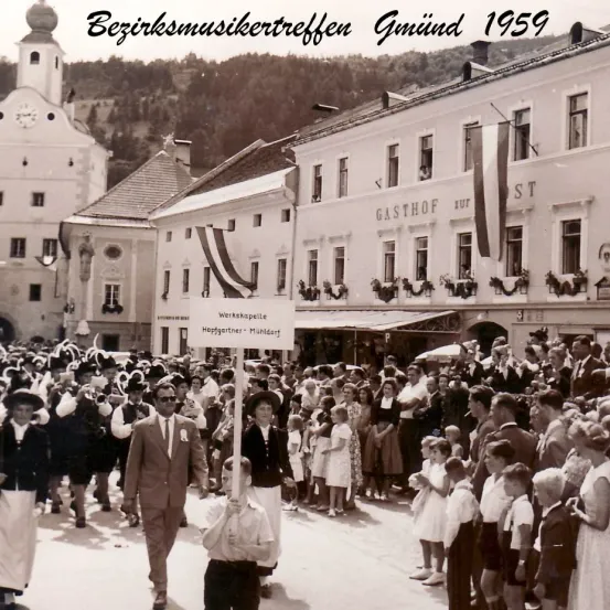Ein altes Schwarz-Weiß-Foto zeigt eine Parade auf einem Stadtplatz. Das Schild lautet Werkskapelle Hopfgartner-Muldorf. Gebäude säumen die Straße, eines mit Gasthof. Menschen in historischer Kleidung versammeln sich, um zuzusehen.