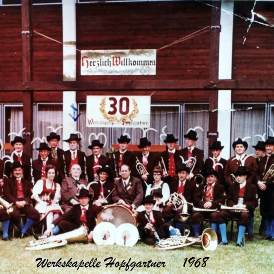 Eine Gruppenaufnahme der Werkskapell Hopfgartner-Band aus dem Jahr 1968, die vor einem Gebäude mit einem Schild steht, auf dem 'Geschlich Willkommen' steht. Sie tragen alle traditionelle Kleidung und halten Musikinstrumente in den Händen.