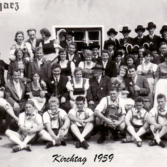 Ein Schwarz-Weiß-Foto von 1959, das eine große Gruppe von Menschen in traditioneller Kleidung zeigt, vermutlich bei einem Kirchenfest. Die Gruppe sitzt und steht vor einem Gebäude mit einem Schild, das 'Marc3' liest.