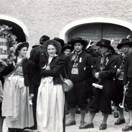Ein Schwarz-Weiß-Foto einer Gruppe von Menschen, einschließlich Musiker, die vor einem Gebäude stehen, in traditioneller Kleidung und mit Hüten. Eine Frau lächelt in der Mitte.