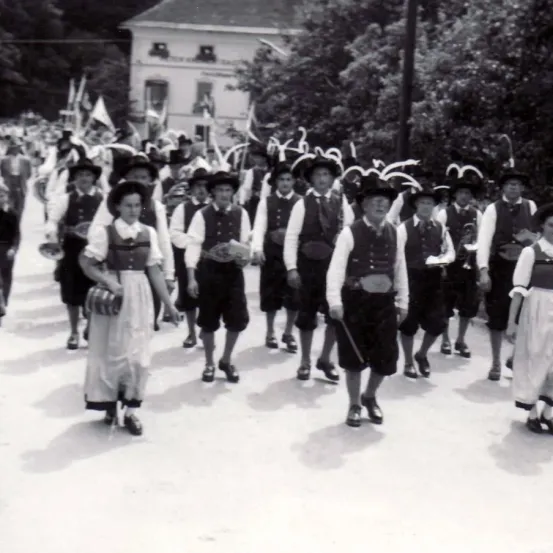 Ein Schwarz-Weiß-Foto zeigt eine Gruppe von Menschen in traditionellen Kostümen, die bei einer Parade marschieren. Einige halten Musikinstrumente und scheinen in einer Reihe zu gehen.