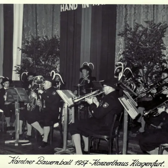 Ein Schwarz-Weiß-Foto einer Band, die vor einer Bühne mit Pflanzen und einem Textüberlagerung Instrumente spielt.