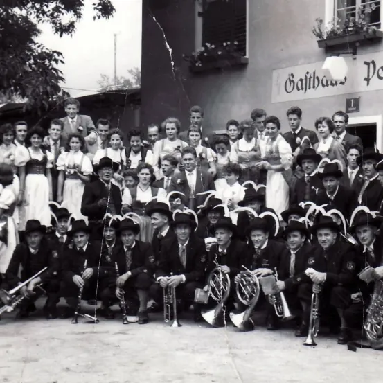 Ein Schwarz-Weiß-Foto einer Gruppe von Menschen in traditioneller Kleidung und Hüten, einige mit Musikinstrumenten, versammelt vor einem Gebäude mit dem Schild 'Gajtbauer'.