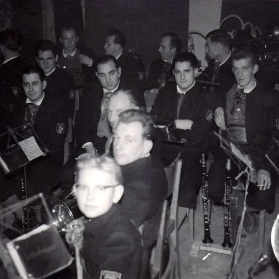 Ein Schwarz-Weiß-Foto einer Gruppe von Männern in Anzügen, die zusammensitzen, möglicherweise ein Orchester oder eine Band, mit Instrumenten wie Trompeten und Klarinetten auf einer Bühne.