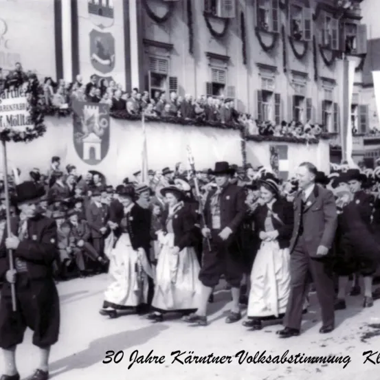 Ein Schwarz-Weiß-Foto zeigt eine Parade, bei der Menschen in traditioneller Kleidung vor einem Gebäude marschieren. Zuschauer beobachten von Balkonen und Gehwegen. Das Foto ist mit '30 Jahre Kärntner Volksabstimmung KL' beschriftet.