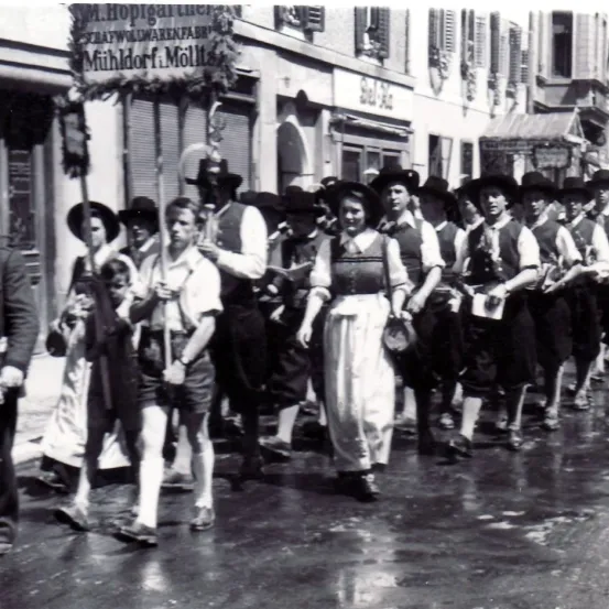 Eine Gruppe von Menschen in traditioneller Kleidung geht eine Straße hinunter vor einem Gebäude mit einem Schild, das 'Mühldorf: Möllt' liest.