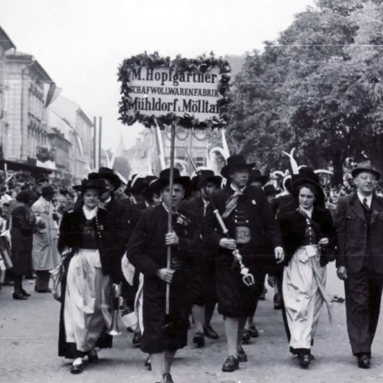 Ein altes Foto einer Parade in Mühldorf, Molltar. Teilnehmer in traditioneller Kleidung, die ein Banner für M. Hopfgartner Schafwollwarenfabrik tragen.