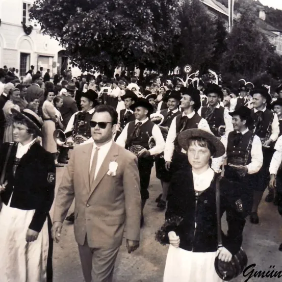 Ein Schwarz-Weiß-Foto einer Parade mit mehreren Personen in traditioneller Kleidung. Einige spielen Musikinstrumente. Der Mann in der Mitte trägt eine Sonnenbrille und einen Anzug.