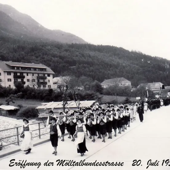 Ein Schwarz-Weiß-Foto zeigt eine Blaskapelle in traditioneller Kleidung, die eine Straße entlangzieht. Das Bild ist vom 20. Juli 1915 datiert.