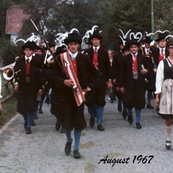 Eine Gruppe von Männern in traditioneller bayerischer Tracht, die im August 1967 bei einem Umzug mitmarschieren. Sie tragen Musikinstrumente und Hüte.