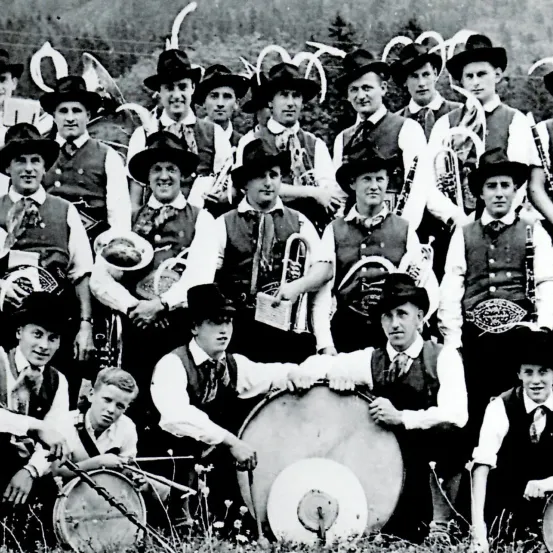 Eine Gruppe von Musikern in traditioneller Kleidung posiert für ein Foto. Sie halten verschiedene Instrumente wie Trompeten, Posaunen und Trommeln. Einige sitzen, während andere hinter ihnen stehen.