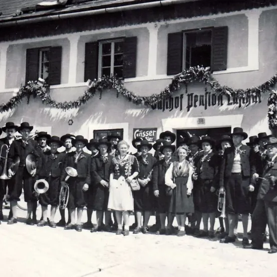 Eine Gruppe von Musikern und Personen in traditioneller Kleidung steht vor einem Gebäude. Das Gebäude ist mit festlichen Dekorationen über dem Eingang verziert.