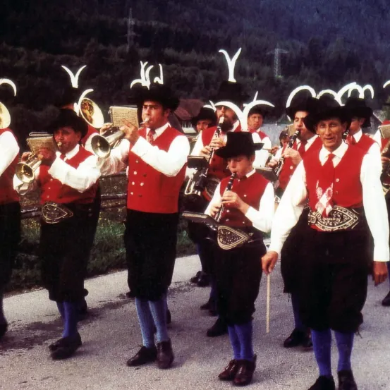 Eine Gruppe von Musikern in traditioneller Kleidung, einschließlich roter Westen, schwarzer Hose und Hüten, spielt auf der Straße. Sie halten verschiedene Instrumente, darunter Trompeten, Posaunen und Klarinetten. Im Hintergrund befinden sich Berge und ein Turm.