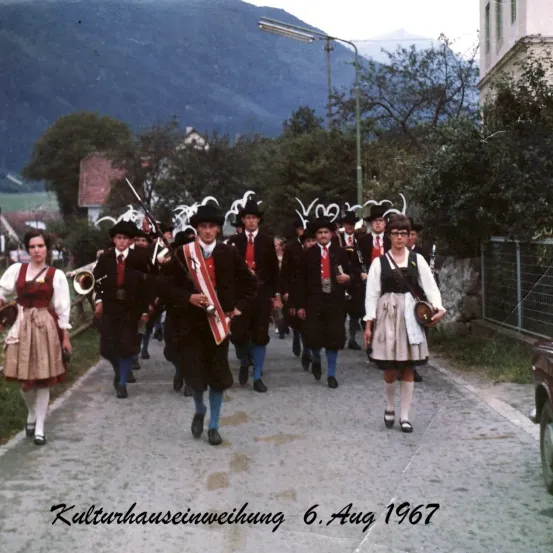 Eine Gruppe von Menschen in traditioneller Kleidung, die wahrscheinlich für eine kulturelle Veranstaltung eine Straße hinuntermarschiert, mit Bergen im Hintergrund am 6. August 1967.