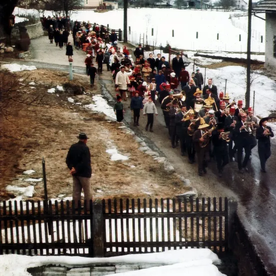 Eine große Menschenmenge marschiert einen verschneiten Pfad hinunter, mit einer Blaskapelle, die vor ihnen Musikinstrumente spielt. Die Menge trägt Winterkleidung.