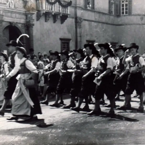 Eine Gruppe von Menschen in traditioneller Kleidung, die bei einer Parade, vermutlich für ein Fest, marschieren. Die Leute tragen Hüte und halten Instrumente. Die Menge hinter ihnen scheint zuzuschauen.