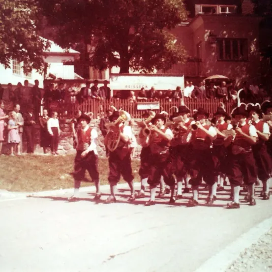 Eine Gruppe von Musikern in traditioneller Kleidung marschiert auf einer Straße, mit Zuschauern an der Seite.