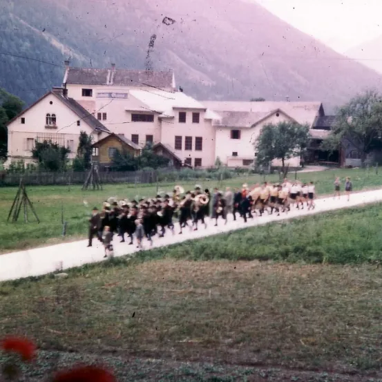 Eine Gruppe von Menschen in Uniformen marschiert auf einem Pfad in einer ländlichen Gegend mit Bergen im Hintergrund.