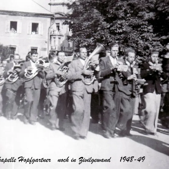 Ein Schwarz-Weiß-Foto einer Marschkapelle mit der Bildunterschrift 'kapelle Hopfgartner nach in Zivilgewand 1948-49'. Die Musiker spielen verschiedene Blechblasinstrumente.