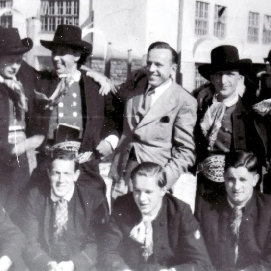 Ein altes Schwarz-Weiß-Foto einer Gruppe von Männern in Anzügen und Hüten, die lächeln, einige mit Krawatten und Gürteln, vor einem Gebäude.