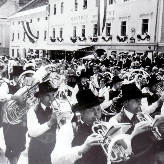 Ein Schwarz-Weiß-Foto zeigt eine Blaskapelle, die auf einem Stadtplatz auftritt. Die Musiker tragen traditionelle Kleidung, einschließlich Hüte und Hosenträger. Dahinter versammelt sich eine Menge, um zuzusehen, mit einem Gebäude im Hintergrund, das mit Gasthof beschriftet ist.