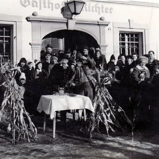 Ein Schwarz-Weiß-Foto zeigt eine Gruppe von Menschen, die vor einem Gebäude versammelt sind. Ein Tisch mit einer weißen Tischdecke und verschiedenen Gegenständen steht in der Mitte. Einige Leute tragen Winterkleidung und Mützen. Eine Lampe hängt über dem Gebäude.