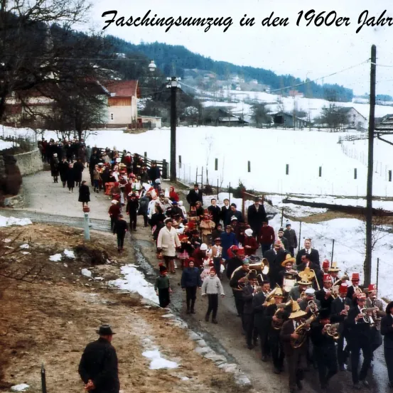Ein Foto aus den 1960er Jahren zeigt eine Gruppe von Menschen, die mit Blechblasinstrumenten in einem verschneiten Dorf marschieren.