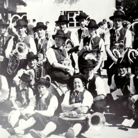 Eine Gruppe von Musikern in traditioneller Kleidung, einige stehend und einige sitzend, posiert für ein Foto. Sie halten verschiedene Blechblasinstrumente.