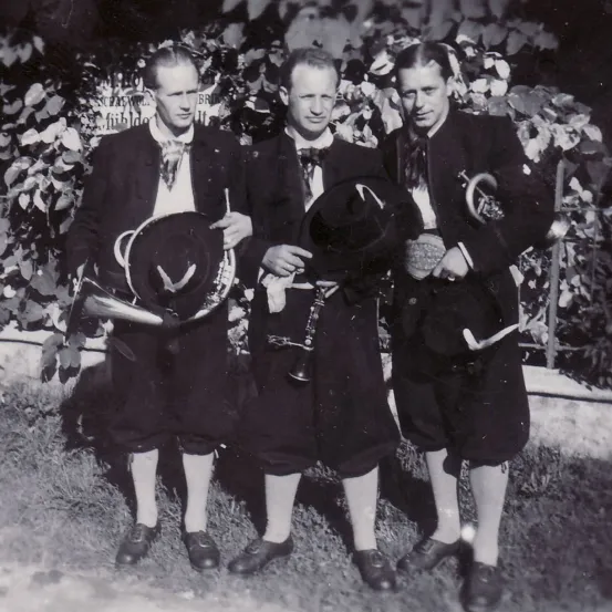 Drei Männer in Anzügen posieren für ein Foto, jeder hält einen Hut und ein Musikinstrument in der Hand und steht vor einem grünen Busch.