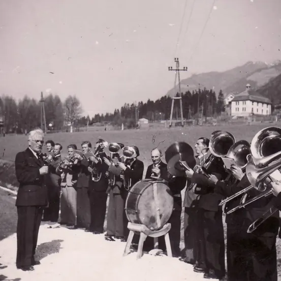 Eine Gruppe von Musikern in Anzügen spielt Blechblasinstrumente im Freien mit einem Gebirgszug im Hintergrund.