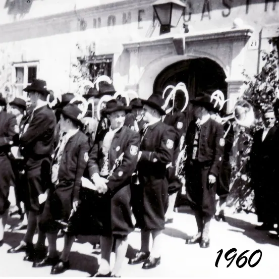 Ein Schwarz-Weiß-Foto aus dem Jahr 1960 zeigt eine Gruppe von Menschen, wahrscheinlich bei einer Parade, in Uniformen, Hüten und Schuhen. Sie stehen vor einem Gebäude mit einer Lampe auf der Spitze.