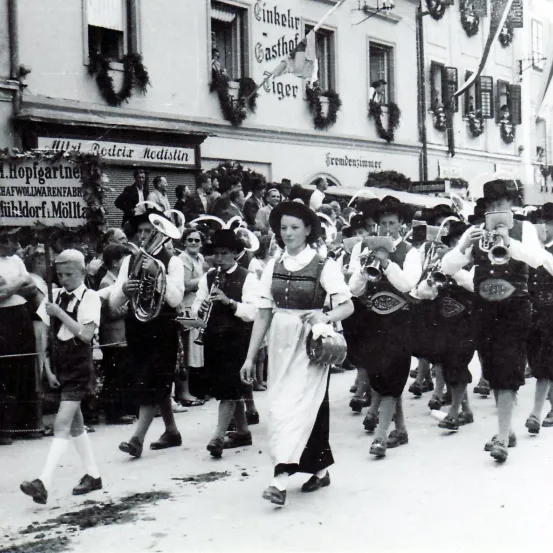 Ein altes Foto zeigt eine Gruppe von Musikern, die in traditioneller Kleidung mit Instrumenten wie Trompeten und Posaunen marschieren. Zuschauer versammeln sich an den Straßenrändern. Die Szene spielt vor einem Gebäude mit festlichen Dekorationen.