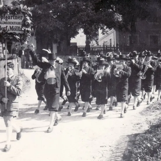 Ein Schwarz-Weiß-Bild zeigt eine Marschkapelle in historischer Kleidung, angeführt von einem Mann in einem Zylinder, die eine Straße entlangzieht, geschmückt mit einem Schild, das 'Splgartner' steht. Die Bandmitglieder spielen verschiedene Instrumente.