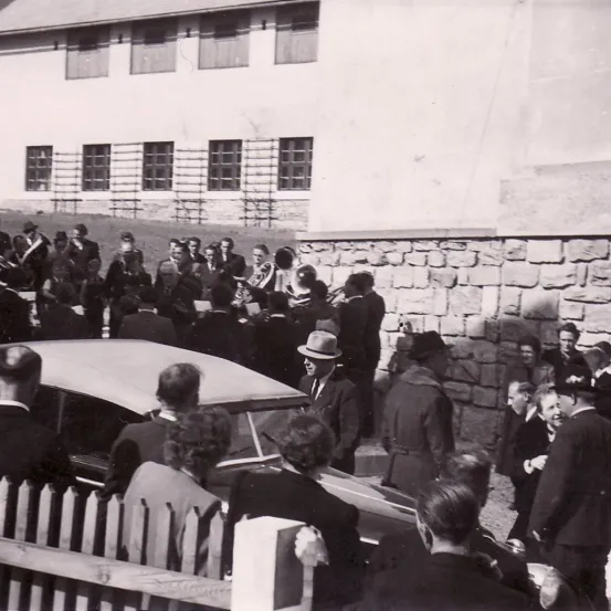 Ein Schwarz-Weiß-Foto zeigt eine Menschenmenge, die sich um ein Auto versammelt hat, möglicherweise bei einer Beerdigung oder Gedenkfeier. Einige Personen sind formell gekleidet, und einige spielen Blechblasinstrumente. Im Hintergrund ist ein Gebäude mit Fenstern und einer Steinmauer zu sehen.