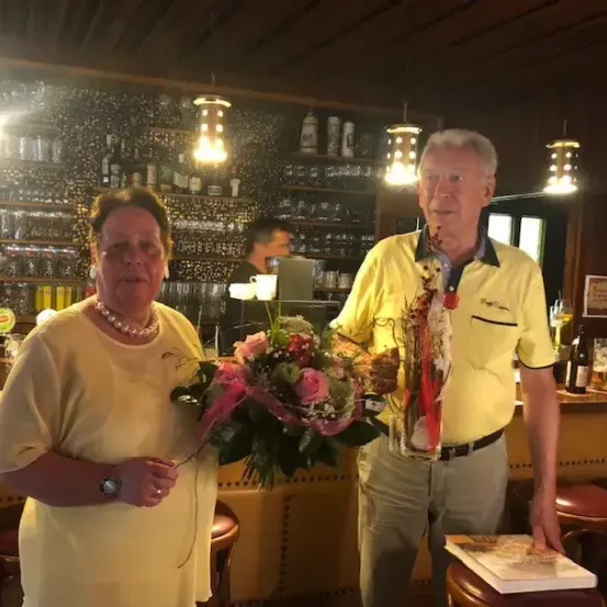 Ein älteres Paar steht in einer Bar, die Frau hält einen Blumenstrauß und der Mann ein Buch und Blumen. Hinter ihnen befinden sich Regale mit Flaschen und Gläsern.