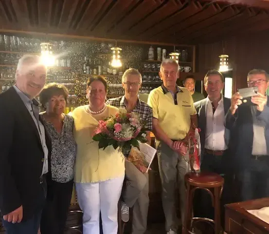 Mehrere Menschen sind zu einer Feier in einer Bar versammelt. Eine Frau hält Blumen, und zwei Männer halten ein Buch und eine Flasche. Dahinter befinden sich Regale mit Flaschen und Gläsern.