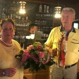 Ein älterer Mann und eine Frau stehen lächelnd in einer Bar. Die Frau hält einen Blumenstrauß, und der Mann hat einen Blumenstrauß. Sie sind beide in Gelb gekleidet. Hinter ihnen steht eine Bartheke mit Flaschen und Gläsern.