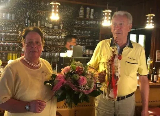 Ein älterer Mann und eine Frau stehen lächelnd in einer Bar. Die Frau hält einen Blumenstrauß, und der Mann hat einen Blumenstrauß. Sie sind beide in Gelb gekleidet. Hinter ihnen steht eine Bartheke mit Flaschen und Gläsern.