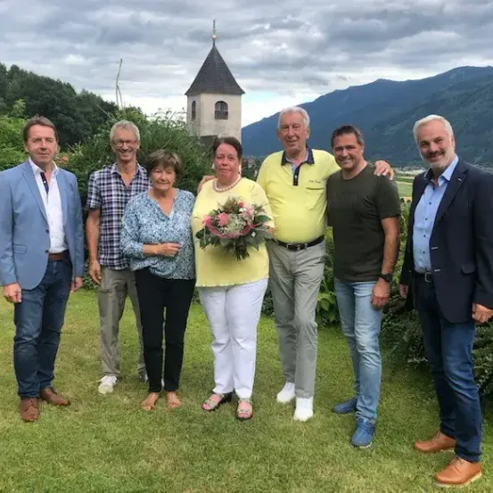 Sechs Personen posieren für ein Foto auf einem grünen Rasen mit Bergen im Hintergrund. Es gibt zwei Frauen und vier Männer. Eine Frau hält einen Blumenstrauß.