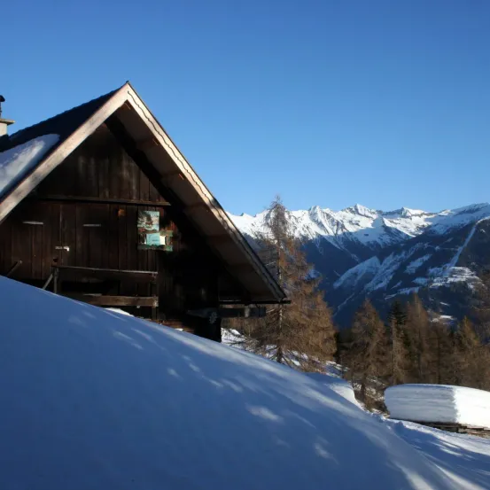 Eine Holzhütte mit schneebedecktem Dach liegt in einer verschneiten Landschaft. Dahinter sind schneebedeckte Berge unter einem klaren blauen Himmel sichtbar.