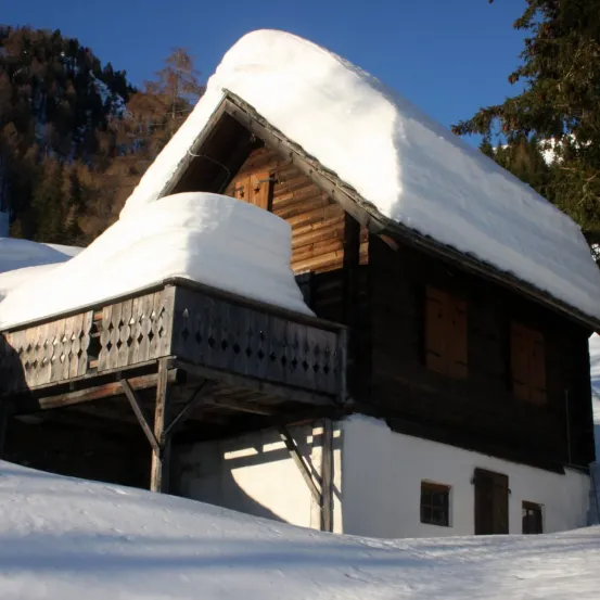 Ein rustikales Holzhaus mit schneebedecktem Dach und Balkon steht in einer verschneiten Landschaft. Das Haus ist von Bäumen und Bergen umgeben.