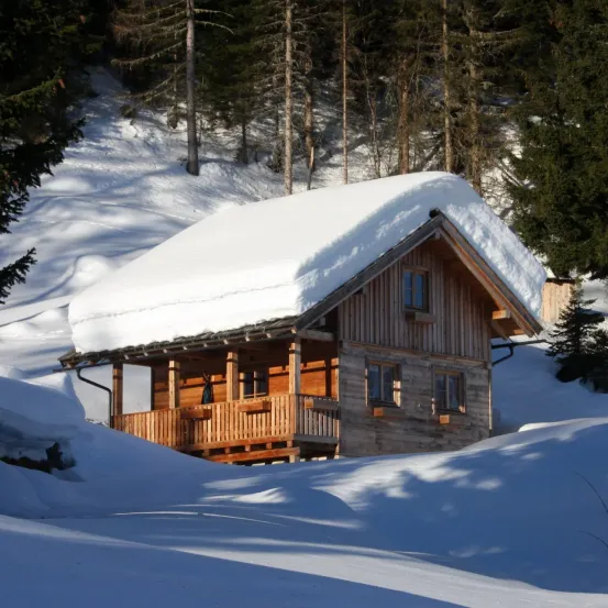 Eine kleine Holzhütte, die von Schnee bedeckt ist, wird von einer verschneiten Landschaft umgeben. Die Hütte hat eine hölzerne Veranda und mehrere Fenster. Schneebedeckte Bäume umgeben die Hütte.