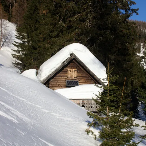 Ein kleines Holzhaus, das von Schnee bedeckt ist, liegt in den Bergen. Ein Solarpanel ist am Dach befestigt. Das Haus ist von Kiefern umgeben.